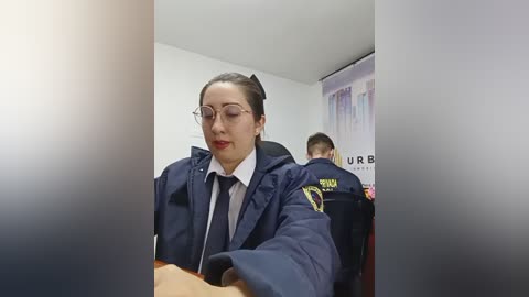 Video of a woman with fair skin, brown hair, and glasses in a navy jacket, seated at a desk. Behind her, two men in dark uniforms with \"URBAN\" logos are visible.