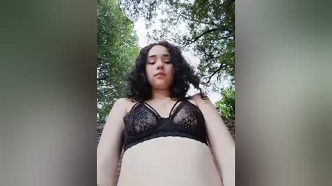 Video of a young woman with curly black hair, wearing a black lace bra, standing in a lush green outdoor setting with trees.