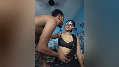 Video of a shirtless, muscular Black man with glasses holding a blindfolded, busty, light-skinned woman in a black bra, seated on a patterned carpet in a dimly lit room with blue walls.