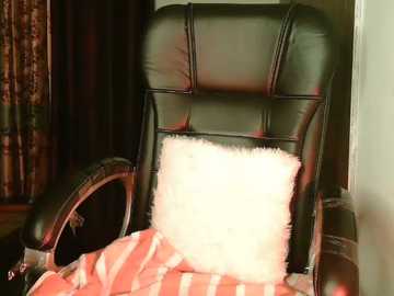 Video of a plush black leather office chair with a white fluffy pillow and a red-striped blanket draped over the backrest, set against dark, patterned curtains in the background.