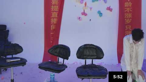 Video of a festive celebration featuring three black plastic chairs in front of red banners with Chinese characters on a white wall, confetti scattered on the floor, and a man in a white suit.