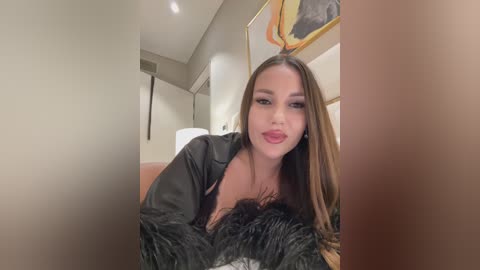 Video of a fair-skinned woman with long brown hair, wearing a black leather jacket and black fur-trimmed jacket, smiling in a modern, warmly lit room with beige walls and abstract artwork.