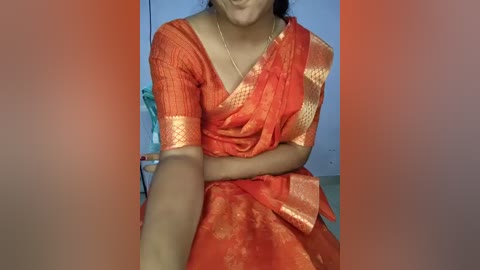 Video of a South Asian woman wearing a vibrant orange saree with gold embroidery, seated indoors, with a blurred background.