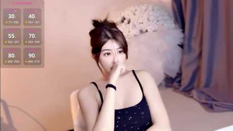 Video of an East Asian woman with fair skin, dark hair in a messy bun, wearing a black polka dot dress, biting her finger, in a cozy, softly lit room with floral decor.