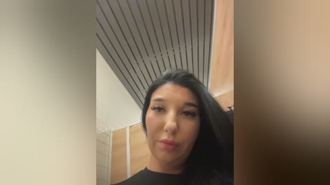 Video of a Latina woman with long black hair, fair skin, and full lips, wearing a black top, in a tiled bathroom with beige walls and a wooden door.
