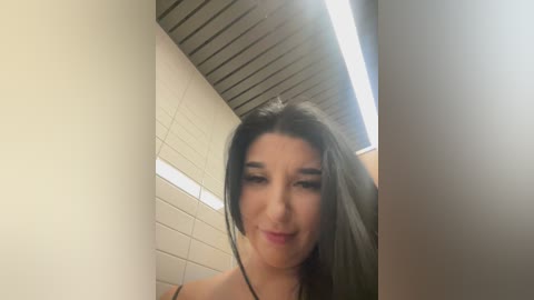 Video of a young woman with long, straight black hair, fair skin, and dark eyes, smiling. She stands in a modern, brightly lit bathroom with white tiled walls and a slanted, wooden ceiling.