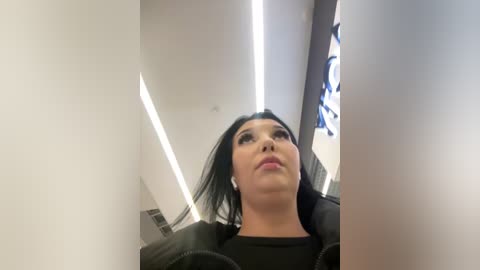 Video of a young woman with straight black hair, wearing a black jacket, standing in a modern, brightly lit elevator. The background includes white walls and a round, blue-patterned clock.