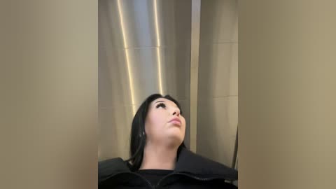 A video of a woman with fair skin and black hair, wearing a black coat, standing in a dimly lit, beige-tiled bathroom with vertical fluorescent lights. She gazes upward, appearing contemplative.