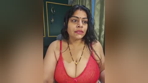 A video of a plus-sized South Asian woman with medium skin tone and long black hair, wearing a red lace bra revealing ample cleavage, and a gold necklace. She sits in a room with green walls and a framed picture in the background.