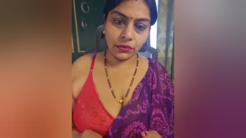 A video of an Indian woman with medium skin tone, dark hair, and a bindi, wearing a red lace bra and a purple sari with floral patterns, standing indoors near a green door and window.