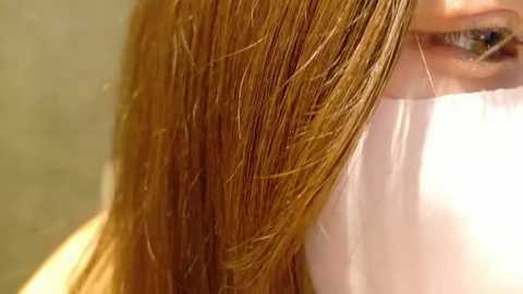 A close-up video of a woman with straight, light brown hair, partially covered by a white surgical mask, revealing her slightly smiling mouth. The background is blurred, focusing on the hair and mask.