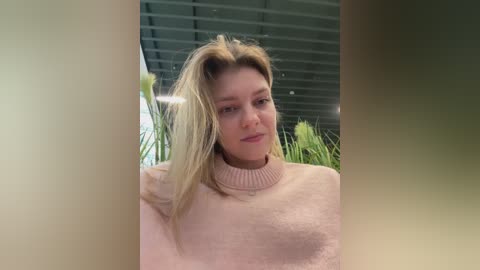 A video of a blonde woman with fair skin, wearing a light pink, fuzzy sweater, standing in front of green foliage under a gray ceiling.