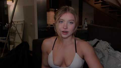Video of a young blonde woman with fair skin, wearing a revealing white bikini top, sitting in a dimly lit living room with a staircase and kitchen in the background.