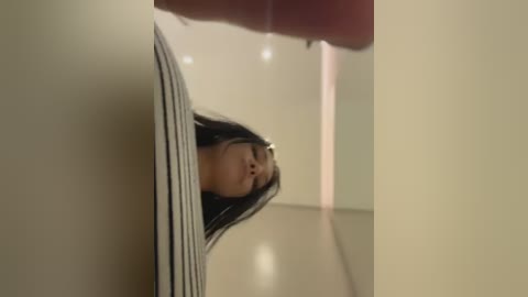 Video of a young Asian woman with long black hair, wearing glasses and a striped top, peering through a narrow gap in a wall.