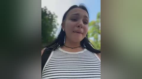 Video of a young woman with long black hair, wearing a white and black striped tank top, standing outdoors with blurred green trees and blue sky in the background.