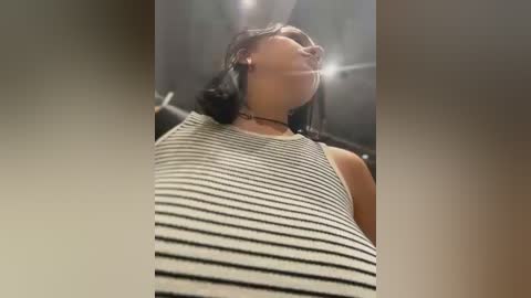 Video of a woman with straight black hair, wearing a sleeveless, striped top, standing against a blurred background with a bright spotlight.