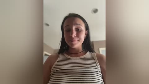 A video of a young woman with long black hair, wearing a striped tank top, standing indoors with neutral beige walls and ceiling lights.