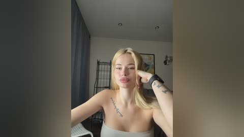 Video of a blonde woman with fair skin and a tattoo on her collarbone, posing with a hand on her cheek, in a minimalist bedroom with grey curtains and a black metal shelf.