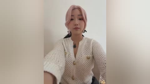 A video of a young Asian woman with light skin and pink hair, wearing a cream-colored knitted cardigan and black pants, captured from a low angle, standing in a narrow, beige-walled hallway.