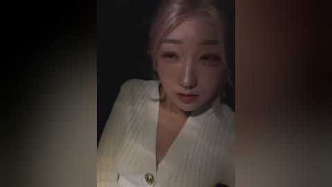 Media: Video of a young East Asian woman with light skin and pink hair, wearing a white, textured cardigan. She has a neutral expression. Background is dark and blurred.