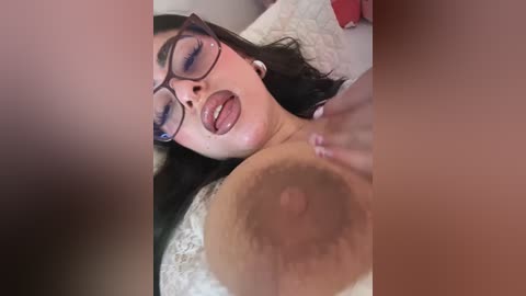 Video of a woman with glasses and black hair, lying on a bed, with a man's hand fondling her large, dark areola.