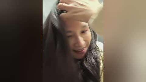 Video of a young Asian woman with long black hair, wearing a yellow top, touching her forehead with her right hand. She has a concerned expression. Background is blurred, with muted colors.