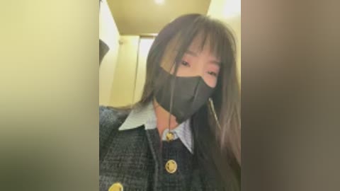 Video of an Asian woman with long black hair, wearing a black face mask, a dark grey tweed blazer, and a white shirt with a bow tie. She is in a dimly lit hallway with beige walls.
