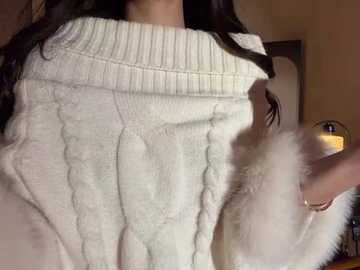 A video captures a person wearing a white, textured cable-knit sweater, holding a fluffy white cat with a pink nose, in a dimly lit room.