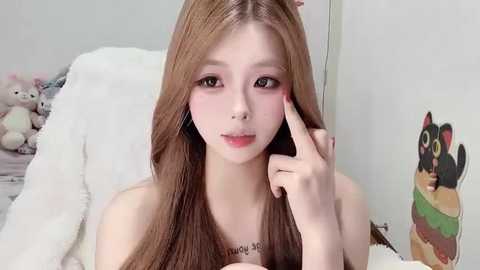 Video of a young East Asian woman with long brown hair, fair skin, and light makeup, sitting on a white bed, touching her face, surrounded by stuffed toys and a cartoon illustration on the wall.