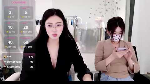 A video depicts two East Asian women in an office. One wears a black blouse, the other a beige sweater, both holding phones. A digital screen displays health data.