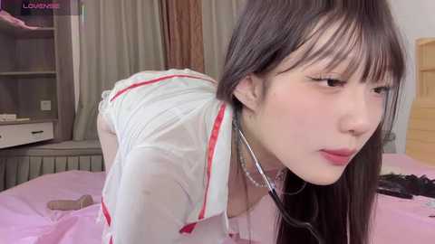 Video of an Asian woman with long, straight black hair, wearing a white nurse costume, bent over a bed, with a stethoscope around her neck, in a bedroom.