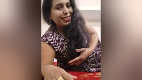 Video of a South Asian woman with long black hair, wearing a maroon sari with white floral patterns, smiling warmly, sitting on a red-patterned cushion.
