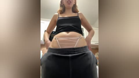 Video of a young woman with light skin and medium-length brown hair, wearing a black tank top and loose black sweatpants, revealing pink panties. She stands confidently indoors with a neutral background.