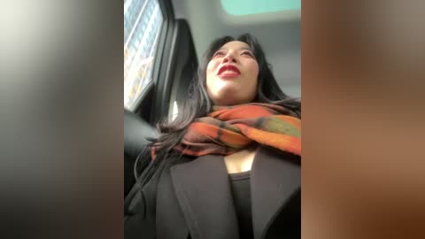A video of a woman with long black hair, wearing a black coat and a colorful scarf, looking up with a slight smile, inside a dimly lit car with a window in the background.