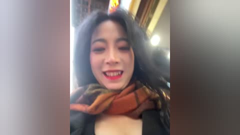 Video of a smiling woman with long black hair, wearing a black top and a plaid scarf, indoors with blurred background.