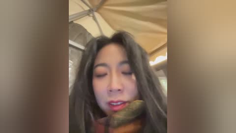A video of an Asian woman with long black hair, wearing a red scarf and closed eyes, standing under a beige tent in a blurred outdoor setting.