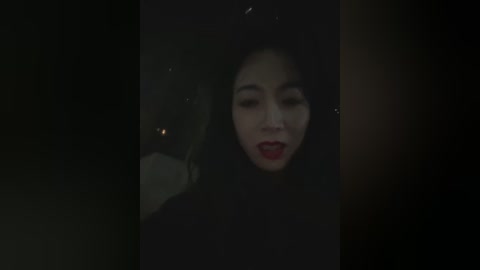 A dimly-lit video of a young Asian woman with long black hair and red lipstick, wearing a dark coat, looking up with a distressed expression. The background is blurred and dark, enhancing the somber mood.