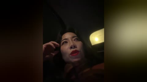 Video of a woman with dark hair and red lipstick, wearing a dark top, looking pensive in dim lighting, with a bright lamp in the background.