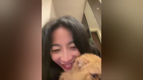Video of a joyful young woman with long black hair, smiling widely, embracing a fluffy orange cat in a warmly lit indoor setting with soft, blurred background.