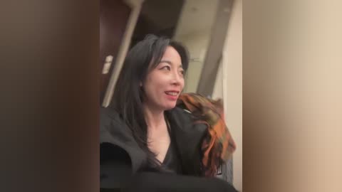 Video of a smiling woman with long black hair, wearing a black dress, holding a plaid scarf. She is indoors, in a hallway, with a blurred background.