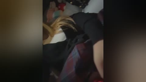 Video of a woman with blonde hair lying on a bed, wearing a black shirt and red plaid skirt, blurred background.
