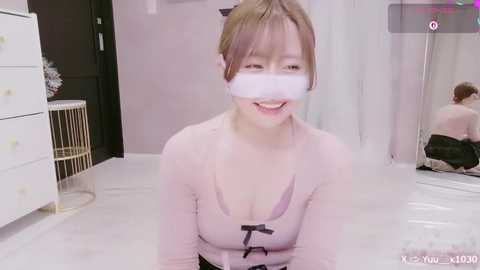 Video of a smiling, light-skinned Asian woman with brown hair, wearing a pink top and white blindfold, kneeling on a white floor in a minimalist room with white furniture and a mirror.