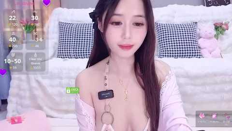Video of an East Asian woman with long black hair, fair skin, and red lipstick, wearing a white lace top, sitting on a white bed with black and white checkered pillows, in a cozy bedroom.