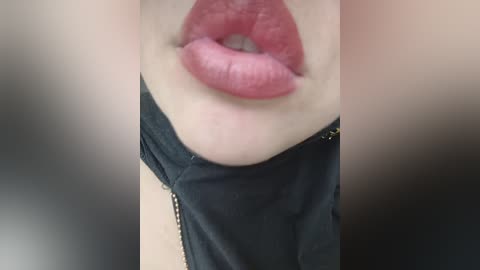Close-up video of a person's mouth with full, glossy pink lips partially covered by a black garment, possibly a jacket. The background is blurry, focusing attention on the lips.