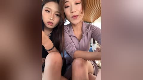 Video of two Asian women, one in a black top and the other in a lavender shirt, sitting close together, smiling, in a room with wooden ceiling panels and a blue bag in the background.