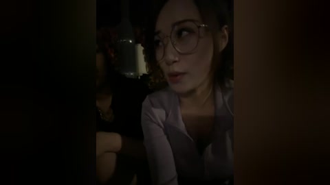 A dimly lit video of a young woman with curly hair, wearing glasses and a light-colored shirt, in a dimly lit room, possibly a bar.