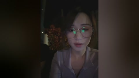 A dimly lit video captures a young Asian woman with curly brown hair and fair skin, wearing large glasses and a light grey shirt. She sits in a dark car, partially visible through the window.