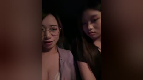 Video of two young Asian women with light skin tones, one wearing glasses and a purple blazer, the other in a black top, set against a dark, possibly nighttime background.
