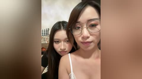 Video of two East Asian women with long black hair, one wearing glasses and a white lace top, the other in a black dress, indoors with a blurred background.