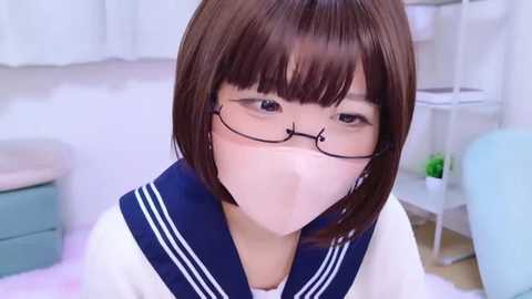 Video of an Asian woman with short brown hair, glasses, and a navy-blue sailor collar shirt, wearing a white mask indoors, blurred background of white furniture and green plants.